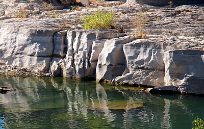 [This rock ledge 6-8 feet high has segments half-way and most of the way up from the water level which are different colors as if the water eroded the rock there.]